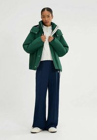 Groene pufferjas met hoge kraag, gedragen over een witte top met rits, gecombineerd met marineblauwe brede broekspijpen en witte sneakers.