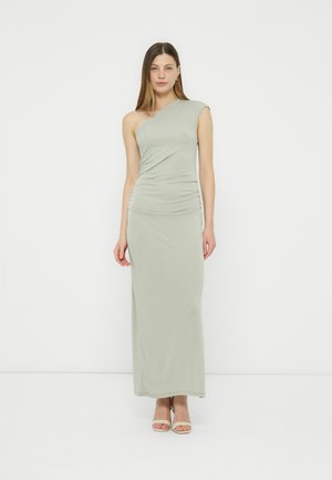 Femeie purtând o rochie lungă de podea, fără mâneci, cu un umăr, verde pal, și pantofi cu toc deschis bej, stând în fața unui fundal alb simplu.