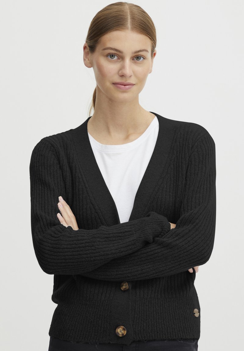 Čierny rebrovaný kardigan s výstrihom do V, predným zapínaním na gombíky a dlhými rukávmi; textúrovaná tkanina pridáva hĺbku. Nosený cez bielu košeľu.
