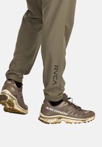 Bruine sportschoenen met gestructureerde zolen, gecombineerd met olijfgroene joggers met een zwart RVCA-logo, elastische boorden en een zachte afwerking.