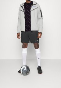 Sudadera gris con cierre, camiseta negra con patrón morado, pantalones cortos negros, calcetines blancos y botas negras. Sostenido un balón de fútbol gris.