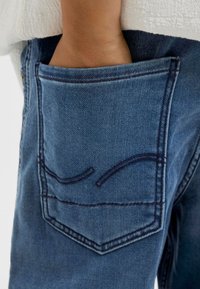 Jeans in denim blu con una grande tasca posteriore caratterizzata da dettagli di cucitura curvi. Il tessuto presenta una texture morbida e un colore sbiadito.