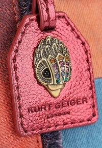 Etichetta in pelle rosa testurizzata con un elemento decorativo dorato che presenta pietre colorate, con il nome del marchio in rilievo "Kurt Geiger, Londra".