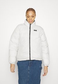 White insulated puffer jacket with high collar, black zipper, and logo detail on chest. Worn over a blue denim skirt. Soft, quilted texture.