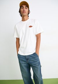 Hvid bomulds t-shirt med et brunt logo på brystet, parret med blå denim jeans med subtile lodrette striber. Modellen bærer en sandfarvet kasket.