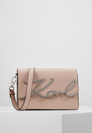 Sac bandoulière rectangulaire en cuir beige avec détail en métal argenté « Karl » et sangle ajustable, présenté sur une surface blanche.