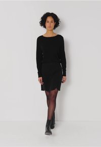 Zwarte trui met een ronde halslijn, gecombineerd met een zwarte rok met een scalloped zoom. Het model draagt donkere panty’s en enkellaarsjes.