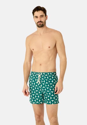 Paitaton mies, jolla on lyhyt parta ja vihreät, valkoisella nauhalla varustetut uimapuvut, seisomassa yksinkertaista vaaleaa taustaa vasten.