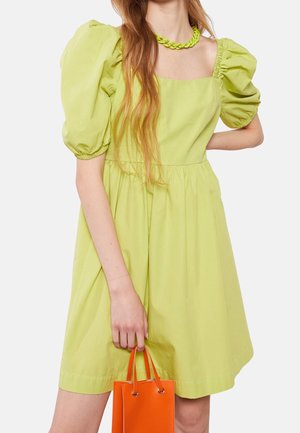 Kvinde iført limegrøn kjole med puffærmer og matchende chunky halskæde, holder en lille, knaldrød-orange håndtaske ved sin side.