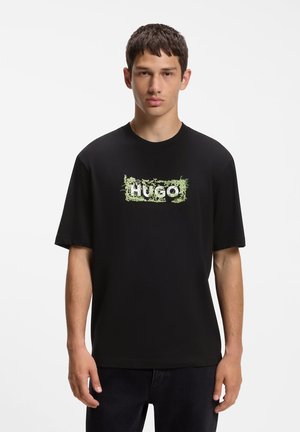 Ung mand iført en sort t-shirt med grønt bladmønster, der danner ordet HUGO på brystet, stående mod en hvid baggrund.