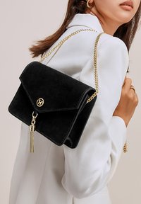 Borsa a tracolla in suede nero con catena dorata, chiusura a patta e un tassello decorativo. Presenta un accento con logo e una texture liscia.