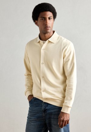Beige overhemd met lange mouwen en kraag, gemaakt van zachte stof. Gecombineerd met blauwe denim jeans, afgebeeld tegen een neutrale achtergrond.