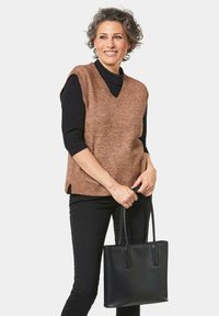 Bruine gebreide vest met een V-hals, gedragen over een zwart longsleeve shirt. Zwarte tote bag in de hand, met zijsplitten op het vest. Zwarte broek.