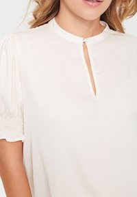Top beige chiaro con scollatura rotonda e apertura a chiave, maniche corte a sbuffo con dettagli arricciati e una consistenza morbida e liscia.