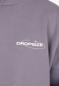 Lila Stoff mit weißem "DROPSIZE"-Textlogo, das ein Orbit-Design und einen Stern an der oberen Brustpartie der Kleidung zeigt.