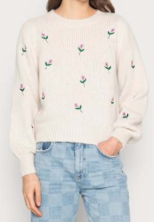 Pull en maille crème avec des fleurs brodées roses, porté avec un jean taille haute bleu clair, une main dans la poche, coupé aux épaules.