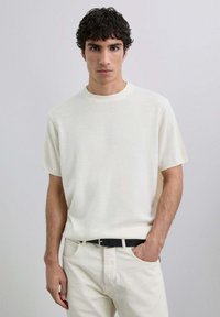Camiseta de mezcla corta en color beige claro, hecha de tejido suave, con cuello redondo y un corte relajado, combinada con pantalones blanco roto y un cinturón negro.