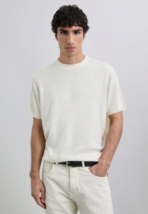 Camiseta de mezcla corta en color beige claro, hecha de tejido suave, con cuello redondo y un corte relajado, combinada con pantalones blanco roto y un cinturón negro.