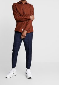 Man med rostfärgad skjorta med knappar, marinblå rutiga byxor, vita Nike-sneakers, står med armarna korsade över torso.