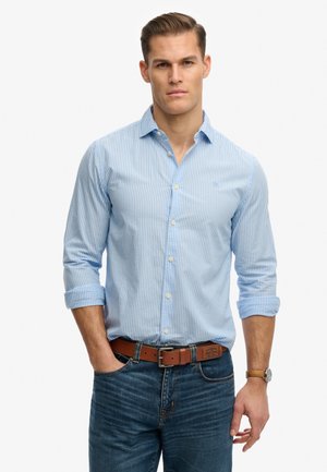 Młody mężczyzna z krótkimi włosami, ubrany w jasnoniebieską koszulę w paski z guzikami, niebieskie dżinsy, brązowy pasek i zegarek na nadgarstku, stoi z jedną ręką w kieszeni.