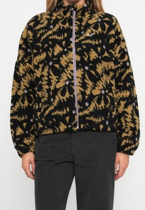 Veste zippée duveteuse avec motif abstrait noir et beige, col montant et poignets élastiques portée avec un pantalon noir.