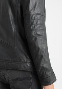 Schwarzer Lederjackenärmel mit abgesteppten horizontalen Linien im Ellenbogenbereich, getragen über dunklen Denim-Jeans.