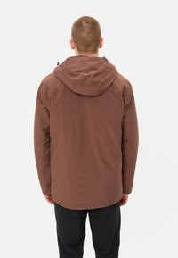 Veste à capuche marron avec une texture lisse et mate. Elle présente une coupe décontractée et des coutures surpiquées, portée sur un pantalon sombre.