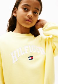 Sudadera de algodón amarilla con cuello redondo, que presenta el texto blanco "HILFIGER" y un pequeño detalle del logo en rojo, blanco y azul.