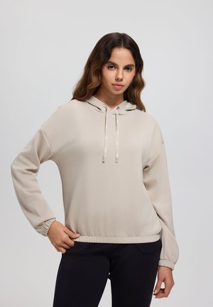 Junge Frau mit welligem Haar, die einen beigen Hoodie mit Kordeln und schwarze Hosen trägt und mit einer Hand in der Hüfte steht.