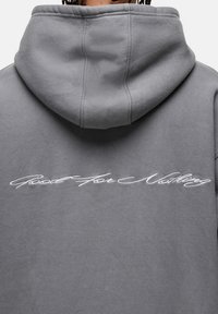 Felpa grigia con cappuccio realizzata in tessuto morbido. Presenta ricami bianchi in corsivo sulla schiena con la scritta "Good For Nothing". Design semplice e rilassato.