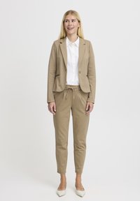 Beige ülikond, millel on hästi istuv bleiser ja paelaga spordipüksid, mille juurde on sobitatud valge kraega särk ja heledat värvi kontsad.