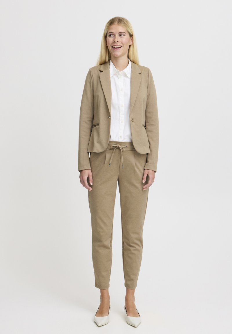 Tenue de costume beige avec un blazer ajusté et un jogging à cordon, associé à une chemise blanche à col et des talons clairs.