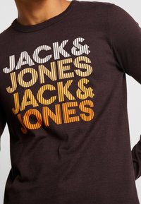 Detail blízko tmavě hnědé košile s dlouhým rukávem, na níž je diagonálně vytištěno "JACK & JONES" třikrát ve sloupcích bílého, žlutého a oranžového textu.