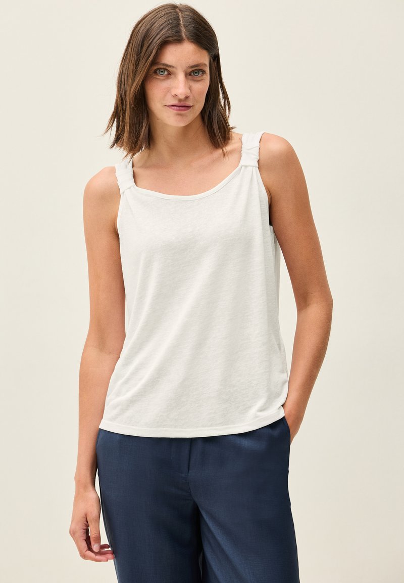 Un haut sans manches blanc en tissu doux présente un col rond et des bretelles torsadées, associé à un pantalon bleu marine.
