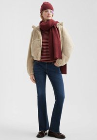 Beżowa, puszysta kurtka na zamek; bordowy, prążkowany szalik; top w maroon i kremowe paski; niebieskie dzwony; brązowe buty; okrągłe okulary.