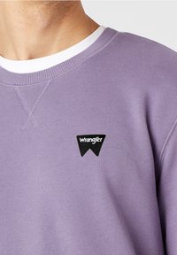 Felpa lavanda realizzata in morbido tessuto, con colletto girocollo a coste bianco e una toppa nera a forma di triangolo con il testo "Wrangler".
