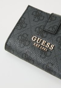 Portefeuille synthétique noir avec une finition texturée, présentant un motif de logo embossé et un matériel doré "GUESS EST 1981" à l'avant.