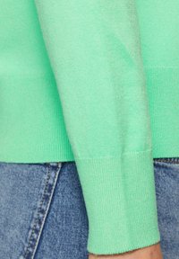 Muntgroene gebreide trui met textuur, featuring ribbel manchetten en onderrand, gecombineerd met denimstof, met een close-up van het armpje.