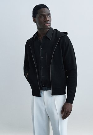 Jeune homme portant un sweat à capuche noir zippé par-dessus une chemise noire à boutons et un pantalon blanc, regardant sur le côté contre un fond uni.