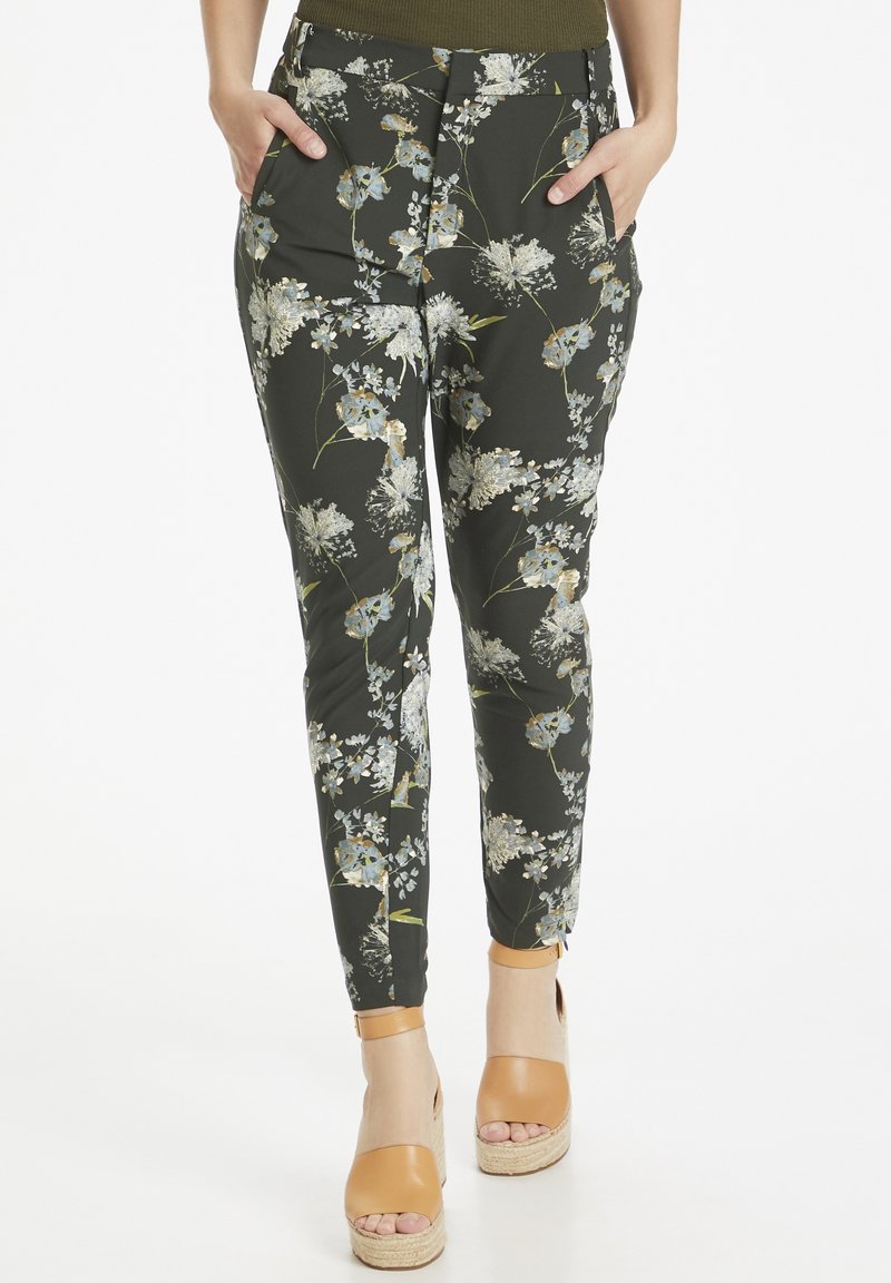 Pantalon à imprimé floral en tissu vert foncé avec des fleurs bleues et blanches. Coupe ajustée, avec poches, porté avec des sandales compensées marron.