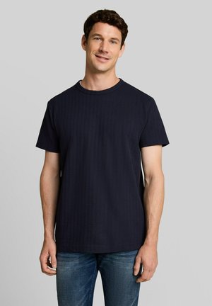 Homme aux cheveux courts, bouclés et bruns, portant un t-shirt noir uni à manches courtes et un jean bleu sur un fond gris clair.