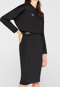 Femme portant un haut noir à manches longues assorti et une jupe de longueur genou avec de petits détails en texte jaune, se tenant avec une main sur la hanche.