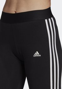 Leggings sportivi neri con una cintura spessa, caratterizzati da tre strisce bianche lungo i lati e un piccolo logo Adidas bianco sul fianco sinistro.