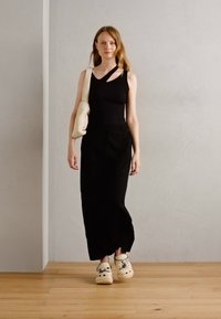 Débardeur noir à côtes, pantalon noir à jambes larges, sac bandoulière crème et sabots blancs à motifs. Fond simple avec un sol en bois.
