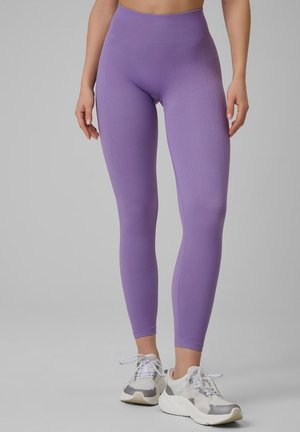 Osoba nosząca wysokie, bezszwowe lawendowe legginsy i białe sportowe buty, stojąca na jednolitym szarym tle.