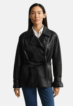 Vrouw met lang donker haar, die een zwart leren double-breasted jas met ceintuur draagt over een wit overhemd en blauwe jeans, staand tegen een witte achtergrond.