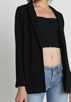 Kobieta ubrana w czarną, otwartą marynarkę, czarny krótki top bez ramiączek i niebieskie dżinsy na jasnym, jednolitym tle.