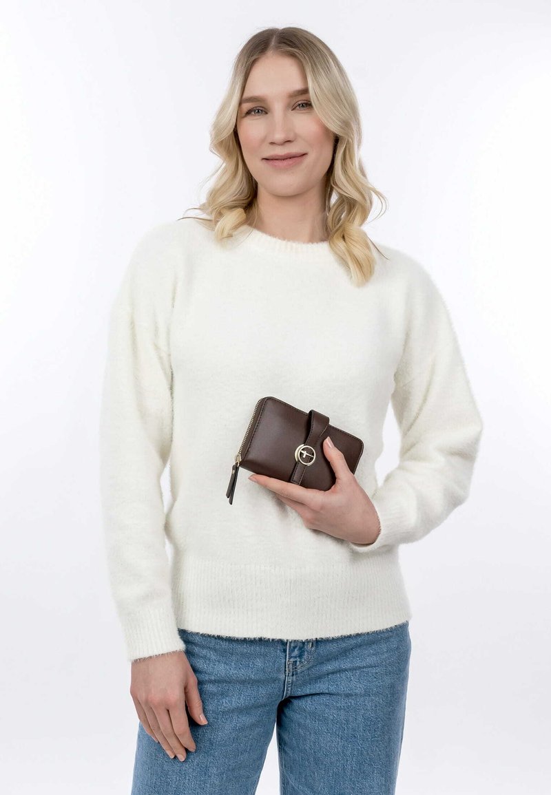 Braune Lederbörse mit einem runden Akzent, in der Hand gehalten. Der Pullover ist weiß und flauschig, dazu blaue Jeans. Einfacher Hintergrund.