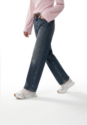 Person iført mørkeblå vide jeans, hvide og pink sneakers, et brunt bælte og en lyserød langærmet top, går fremad.