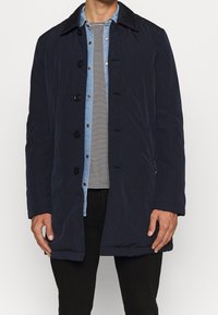 Marineblauer gesteppter Mantel mit Knopfverschluss, kontrastierendem Kragen und Seitentaschen, kombiniert mit einem hellblauen gestreiften Jeanshemd.